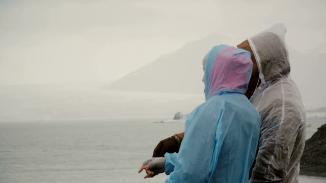 Junges-Paar-in-Regenmäntel-im-Eis-Lagune-in-Island-stehen-und-zeigen-sich-gegenseitig-mit-den-Fingern