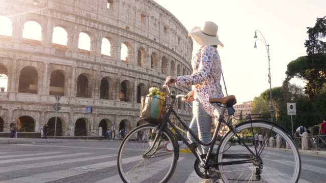 Beautiful-young-woman-in-colorful-fashion-dress-walking-alone-with-bike-crossing-road-in-front-of-colosseum-in-Rome-at-sunset-happy-attractive-tourist-girl-with-straw-hat