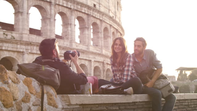 Mochilas-de-tres-turistas-jóvenes-amigos-sentados-mentira-delante-de-Coliseo-de-Roma-al-atardecer-tomando-fotos-fotos-con-dslr-cámara-gafas-de-sol-feliz-hermosa-chica-de-cabello-largo-lenta