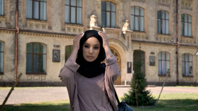 Young-serious-muslim-girl-in-hijab-is-standing-and-fix-her-scarf-in-daytime-in-summer,-watching-at-camera,-building-on-background,-religious-concept