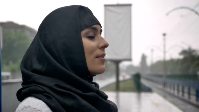 Joven-musulmana-hijab-está-esperando-tren-en-la-estación-de-tren,-lloviendo,-concepto-de-religión,-concepto-urbano.-concepto-de-tiempo,-vista-lateral