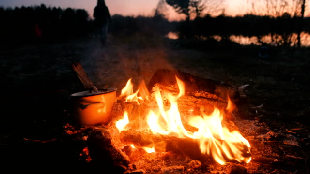 Food-is-cooked-on-the-bonfire-in-a-pan,-open-the-lid-with-branch.-People-in-camping-on-riverbank.