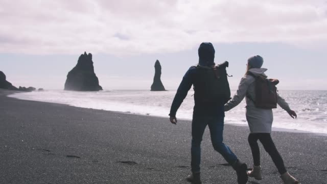 Young-couple-holding-hands-and-running-on-black-sand-beach-in-Iceland,-slow-motion