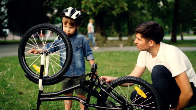 Niño-pequeño-gira-rueda-de-bicicleta-y-pedales-mientras-su-padre-está-hablando-a-él-en-el-césped-en-el-parque-en-día-de-verano.-Familia,-el-ocio-y-el-concepto-de-estilo-de-vida-activo.