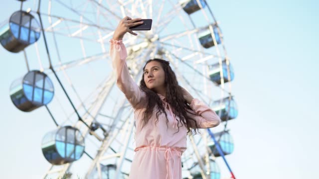 a-girl-with-long-hair-in-a-dress-makes-selfie-using-a-smartphone-standing-near-the-Ferris-wheel.-4K