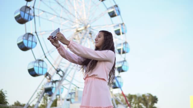 ein-Mädchen-mit-langen-Haaren-in-einem-rosa-langen-Kleid-macht-Selfie-mit-dem-Smartphone-stehen-in-der-Nähe-von-dem-Riesenrad.-4K
