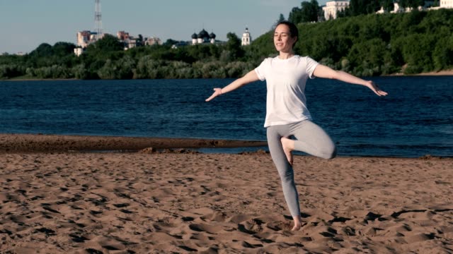 Woman-stretching-yoga-on-the-beach-by-the-river-in-the-city.-Beautiful-city-view.-Vrikshasana-pose.