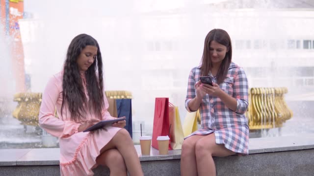 Dos-chicas-después-de-compras-están-sentados-en-el-parque-cerca-de-la-fuente-usando-un-teléfono-y-una-tableta-con-buen-humor.-4K