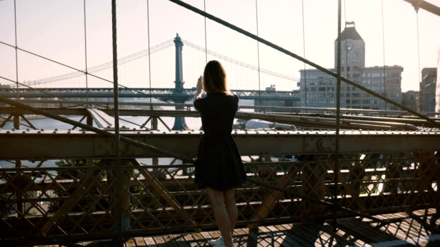 Hermosa-mujer-caucásica-camina-por-el-puente-de-Brooklyn,-Nueva-York,-se-detiene-y-toma-foto-de-smartphone-de-vista-panorámica-4K