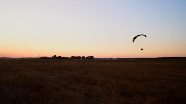 Pilot-flying-a-paraglider-with-a-motor-at-sunset,-the-camera-on-the-suspension-moves-along-the-field