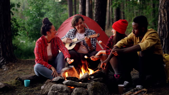 Happy-hikers-are-cooking-marshmallow-on-fire-and-singing-songs-while-handsome-guy-is-playing-the-guitar-during-getaway-in-forest.-Nature,-fun-and-friendship-concept.