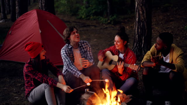 Young-lady-traveler-is-playing-the-guitar,-her-friends-are-cooking-food-on-fire-and-African-American-man-is-clapping-hands.-Friendship,-people-and-music-concept.