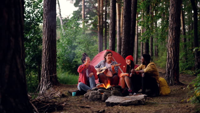 Grupo-multirracial-de-amigos-viajeros-está-cantando-en-el-bosque,-tocando-guitarra-y-comer-melcocha-caliente-está-calentando-alrededor-del-fuego.-Concepto-naturaleza-y-amistad.