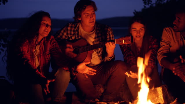 Young-men-and-women-are-singing-songs-to-the-guitar-resting-around-campfire-and-enjoying-music-and-good-company-on-summer-night.-Trees-and-lake-are-visible.