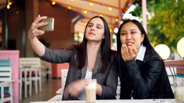 Fröhliche-Freundinnen-machen-Online-Videoanruf-mit-Smartphone-Bildschirm-betrachten-und-sprechen-im-Straßencafé-sitzen-zusammen.-Kommunikation-und-Technologie-Konzept.