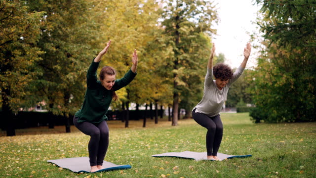 Flexible-girls-are-practising-Chair-position-standing-on-yoga-mats-in-park-and-moving-body-and-head.-Beautiful-autumn-nature,-trees-and-grass-are-visible.
