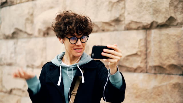 Chica-alegre-está-haciendo-videollamada,-hablando-y-gesticulando-holding-smartphone-y-mirar-pantalla-usando-auriculares.-Concepto-moderno-de-la-tecnología,-la-comunicación-y-la-gente.