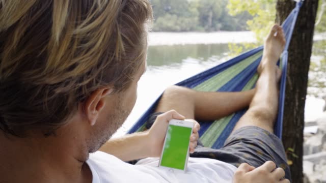 Junger-Mann-mit-Handy-auf-entspannende-Hängematte-am-Fluss-im-Sommer
