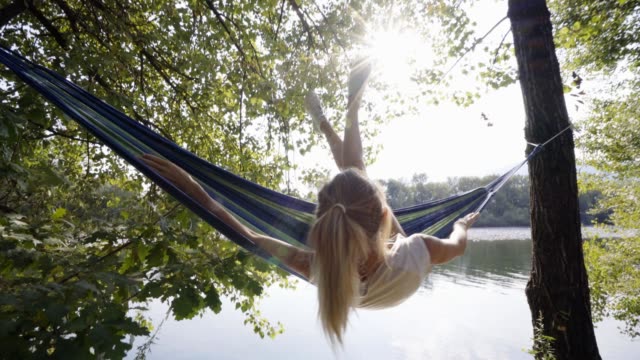 Young-woman-relaxing-on-hammock-by-the-river,-hands-behind-head-enjoying-serene-green-environment.-People-travel-relax-concept-in-vacations