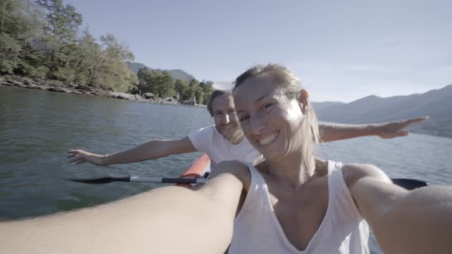 Junges-Paar-in-den-Sommerferien-Kanu-auf-dem-See-in-den-Bergen-zu-genießen.-Die-Leute-Reisen-Spaß-Urlaub-Konzept.-Technologie-und-Jugendkultur-nehmen-Selfie---4K