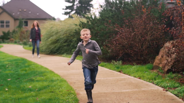 Little-boy-running-down-a-path-at-a-park-while-his-mother-follows,-slow-motion