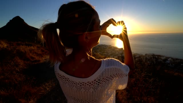 Young-woman-watching-sunset-by-the-sea,-makes-heart-shape