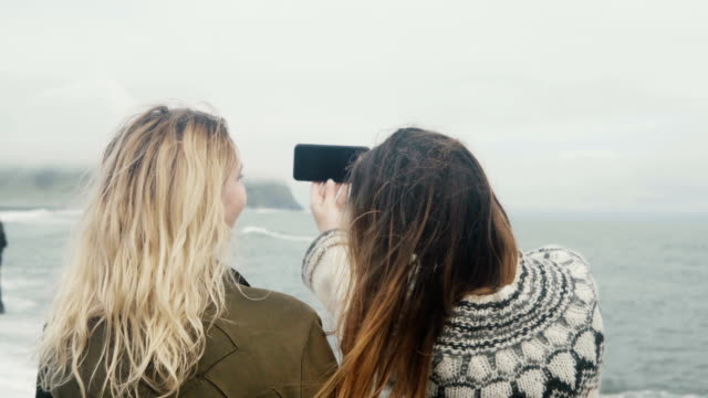 Rückansicht-der-beiden-jungen-Frauen-fotografieren-Selfie-am-Ufer-des-Meeres-in-der-Nähe-der-Troll-Zehen-Felsen-in-Island