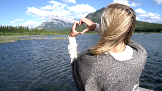 Young-woman-making-heart-shape-finger-frame-on-mountain-lake-scenery