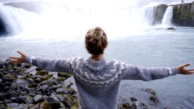 Junge-Frau-vor-dem-herrlichen-Wasserfall-in-Island-ausgestreckten-Armen,-fällt-Godafoss.-Die-Leute-reisen-Exploration-Konzept-4K