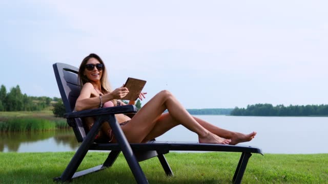 Woman-working-on-a-tablet-computer-traveling-the-world-on-vacation,-sunbathing-on-the-beach-near-the-lake