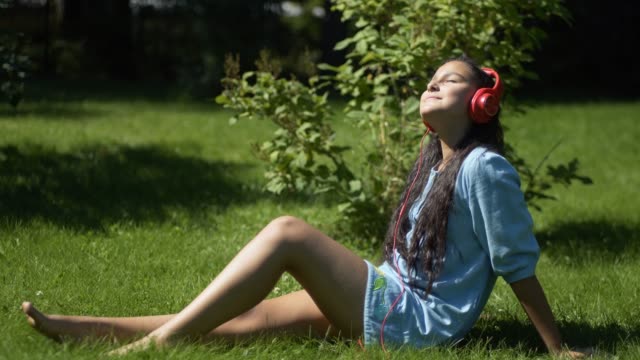 Young-girl-with-long-black-hair-listening-to-music-on-headphones-using-smartphone-sitting-on-grass-in-park-in-sunny-weather.-4K