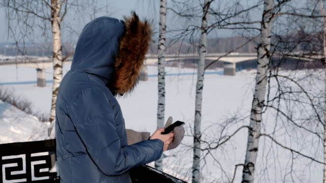 Unrecognizable-woman-in-blue-down-jacket-writes-messaging-in-her-cellphone-in-winter-Park.-Side-view