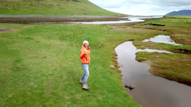Follow-me-to-nature,-girlfriend-waving-hand-to-drone-at-Kirkjufell-mountain-inviting-to-follow-her--People-travel-discovery-concept--4K-aerial-view