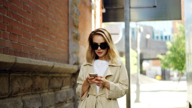 Attraktive-junge-Frau-mit-blonden-Haaren-tragen-von-trendigen-Mantel-und-Sonnenbrille-nutzt-Smartphone-in-modernen-Stadt-zu-Fuß.-Menschen,-moderne-Technologie-und-Jugendkonzept.