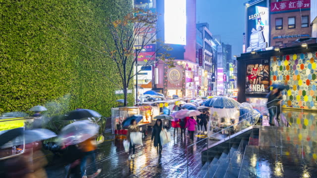 Timelapse-at-Myeong-dong-Market.People-walking-with-umbrellas-on-a-rainy-day-at-shopping-street-at-night,-Seoul,-South-Korea