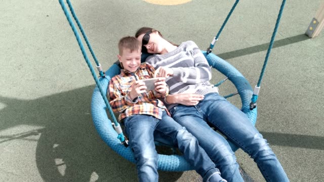 Mom-and-son-swinging-on-empty-webbed-swing-and-playing-game-on-mobile-phone-together.
