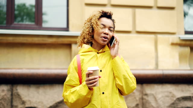 Careless-African-American-girl-is-talking-on-cellphone-and-holding-take-out-coffee-standing-outdoors-in-modern-city.-Communication-and-technology-concept.