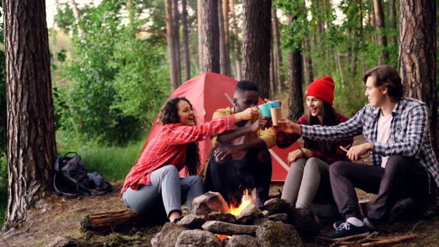 Carefree-tourists-are-toasting,-clinking-glasses-with-hot-drinks-then-drinking-sitting-around-fire-in-forest-near-tent.-Friendship,-recreation-and-youth-concept.