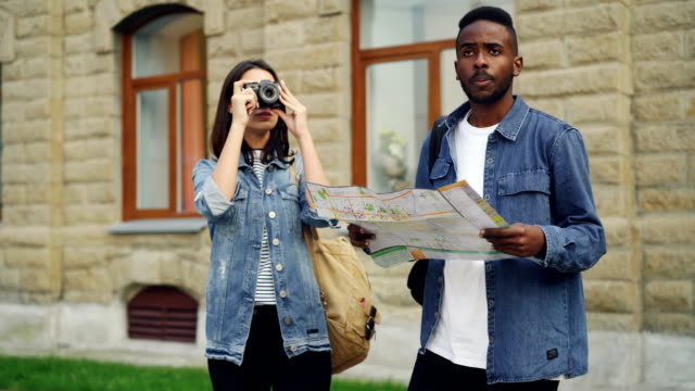 Hübscher-afroamerikanische-Mann-Reisenden-ist-auf-Karte-suchen,-wenn-seine-attraktive-Freundin-Fotos-mit-Kamera-stehend-im-Stadtzentrum-und-im-Gespräch-stattfindet.