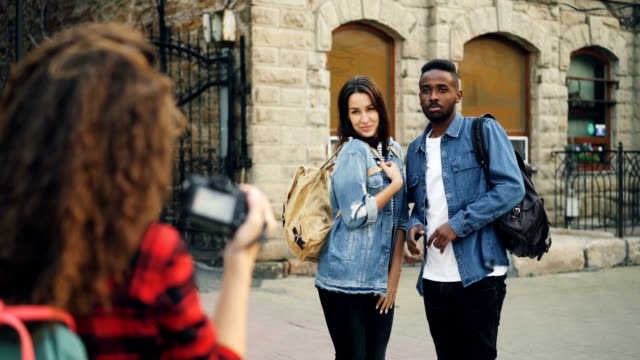 Creative-girl-and-guy-friends-are-posing-for-camera-standing-in-the-street-while-young-woman-with-backpack-is-taking-pictures.-People-are-having-fun-and-laughing.