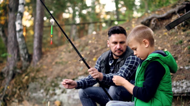 Padre-e-hijo-son-barras-de-explotación-de-pesca-y-hablar,-hombre-enseña-niño-utilizar-equipo-en-día-de-otoño-con-árboles-a-su-alrededor.-Concepto-de-familia,-hobby-y-ocio.
