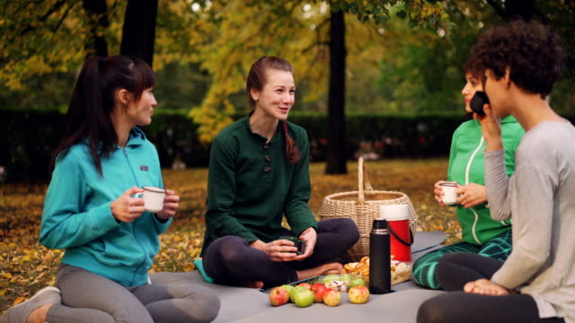 Cheerful-young-women-are-resting-on-mats-having-picnic-talking-and-drinking-tea-after-yoga-class-outdoors-on-beautiful-autumn-day.-Food-and-basket-are-visible.