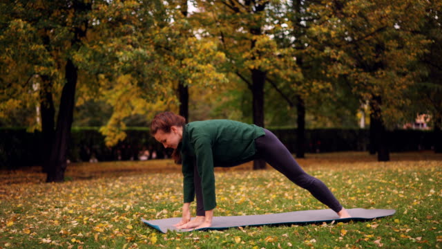 Cheerful-young-woman-is-doing-yoga-balancing-exercises-during-individual-practice-in-city-park-standing-on-mat-and-enjoying-rest-and-nature.-People-and-sports-concept.