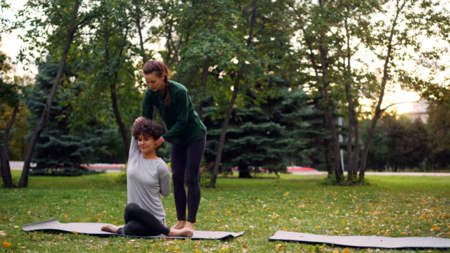Slim-girl-yoga-instructor-is-helping-her-student-to-manage-Gomukhasana-during-individual-practice-in-park-in-autumn.-Guidance,-sports-and-nature-concept.