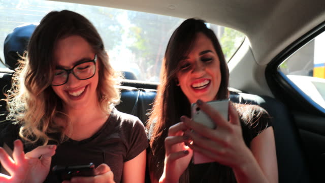 Freundinnen-Surfen-auf-ihrem-Smartphone-während-der-Fahrt-mit-Taxi-zu-lachen.-Frauen-lachen-während-der-Verwendung-von-Handy-auf-dem-Rücksitz-eines-Autos