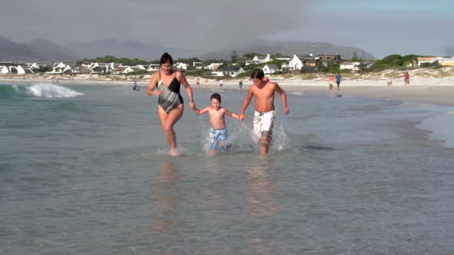 Madre-y-dos-niños-corriendo-a-través-del-agua-superficiales-en-la-playa,-Cape-Town