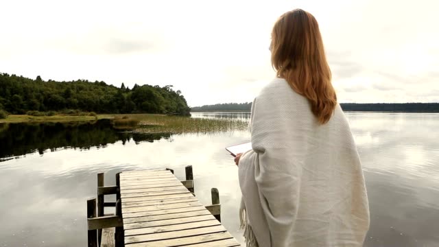 Young-adult-relaxes-on-jetty-above-lake,-uses-digital-tablet