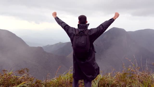 Young-male-hiker-in-raincoat-with-backpack-reaching-up-top-of-mountain-and-raised-hands.-Man-tourist-standing-on-the-edge-of-beautiful-canyon,-victoriously-outstretching-arms-up.-Rear-back-view