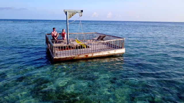 v03848-vuelo-drone-vista-aérea-de-Maldivas-playa-2-personas-pareja-hombre-mujer-relajante-en-la-isla-de-paraíso-tropical-soleado-con-cielo-azul-aqua-agua-mar-4k-flotante-pontoon-embarcadero-el-sol-juntos