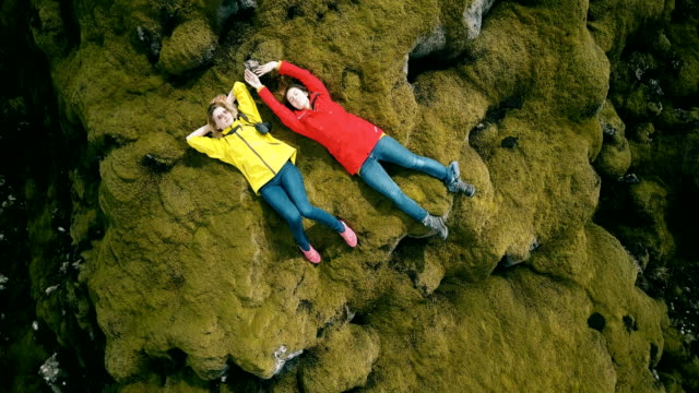 Top-aerial-view-of-two-woman-lying-on-lava-field-in-Iceland-and-rest-after-hiking.-Copter-turning-around-happy-females
